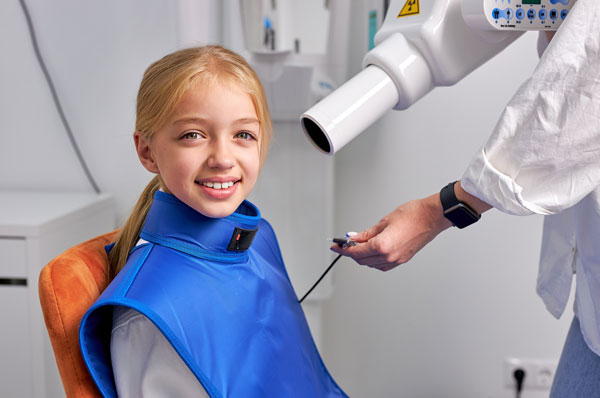 Smiling young girl in dental chair wearing a blue protective lead apron during an X-ray procedure.
