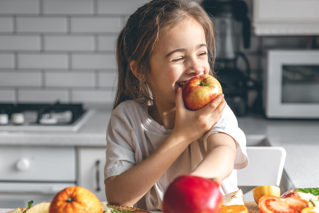 Smiling young girl eating a red apple in a bright kitchen to promote healthy teeth and nutrition